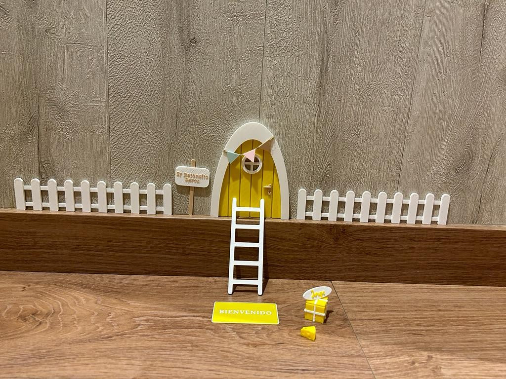 Kit mágico del Ratoncito Pérez con puerta en miniatura amarillo, escalera, valla, cartel de bienvenida y queso, creando un escenario encantador en 3D sobre una superficie de madera.