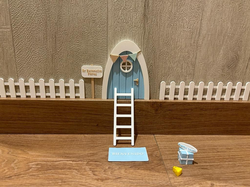 Kit mágico del Ratoncito Pérez con puerta en miniatura azul , escalera, valla, cartel de bienvenida y queso, creando un escenario encantador en 3D sobre una superficie de madera.