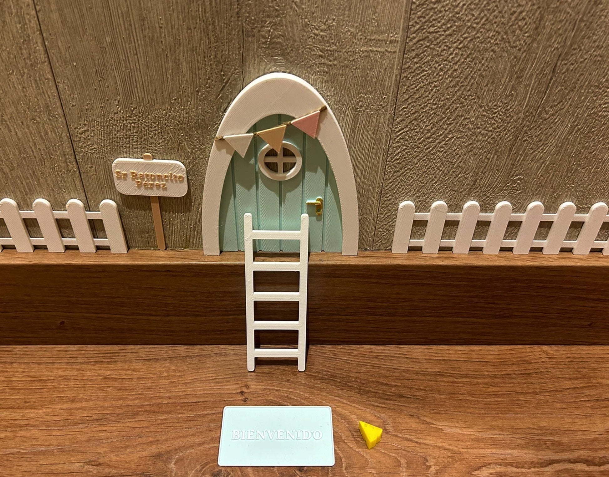 Kit mágico del Ratoncito Pérez con puerta en miniatura, escalera, valla, cartel de bienvenida y queso, creando un escenario encantador en 3D sobre una superficie de madera.